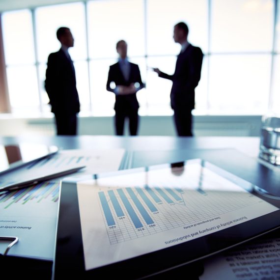 Digital data Close-shot of a tablet computer displaying financial data, three businessmen standing in the background